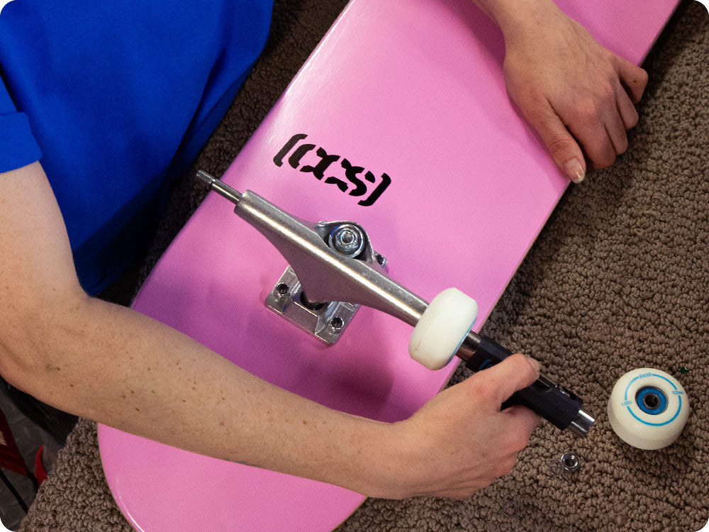 Person building a skateboard, tightening a wheel on a pink board with a handheld skate tool.