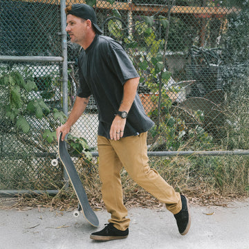 Man wearing CCS Stretch Chinos in Khaki holding a skateboard in an outdoor setting with a chain-link fence and plants in the background.