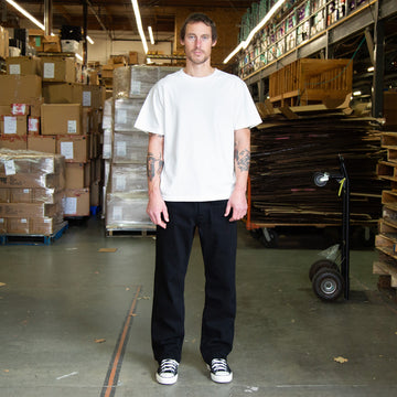 Man standing in a warehouse wearing black jeans and a white t-shirt with stacked boxes and pallets in the background