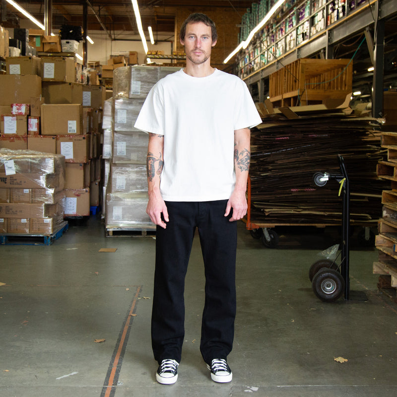 Man standing in a warehouse wearing black jeans and a white t-shirt with stacked boxes and pallets in the background
