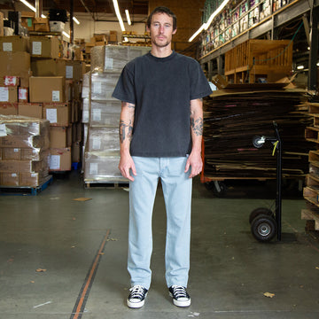 Man standing in a warehouse wearing bleach wash jeans and a faded black t-shirt with stacked boxes and pallets in the background