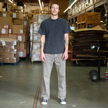 Man standing in a warehouse wearing faded brown jeans and a faded black t-shirt with stacked boxes and pallets in the background