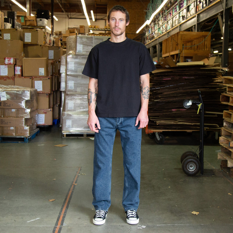 Man wearing a black t-shirt and blue jeans standing in a warehouse with boxes and pallets in the background.